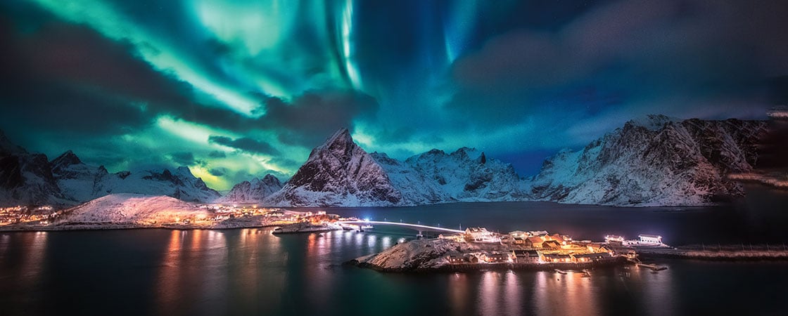 The sky bathed in neon lights with snow covered mountains and city lights below it