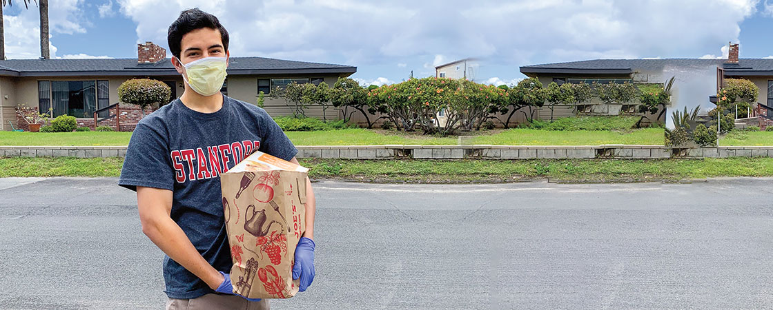 A young man with a mask on standing in front of houses holding a large Trader Joe&apos;s brown bag