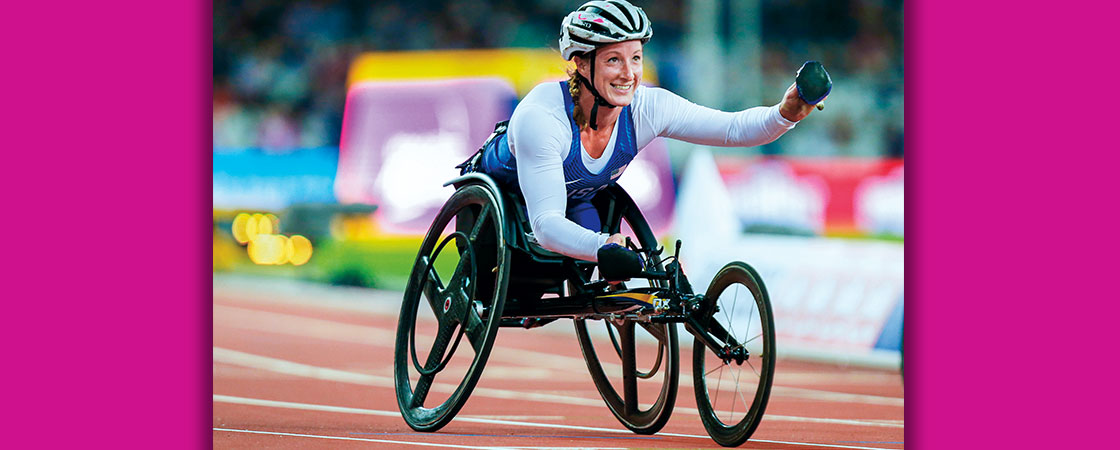 a woman in a racing wheelchair and helmet races in front of a purple background