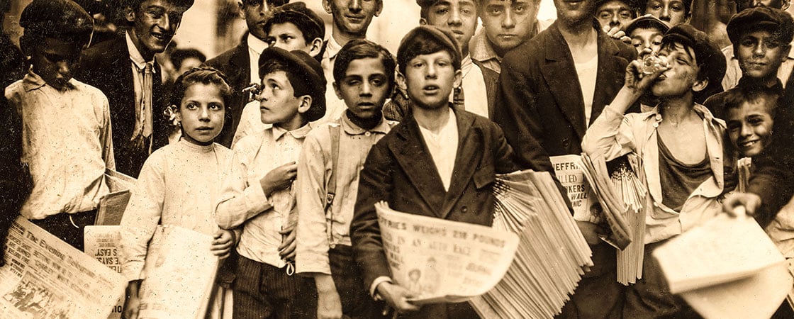 Black and white photo of a group of boys walking and carrying stacks of newspapers