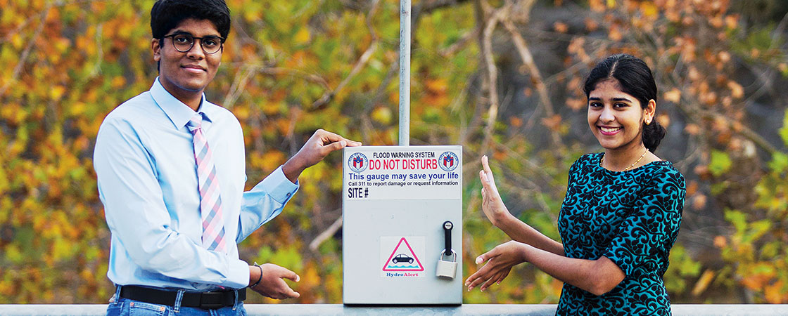 A boy and a girl presenting a metal box  attached to a pole with the text "HydroAlert" on it