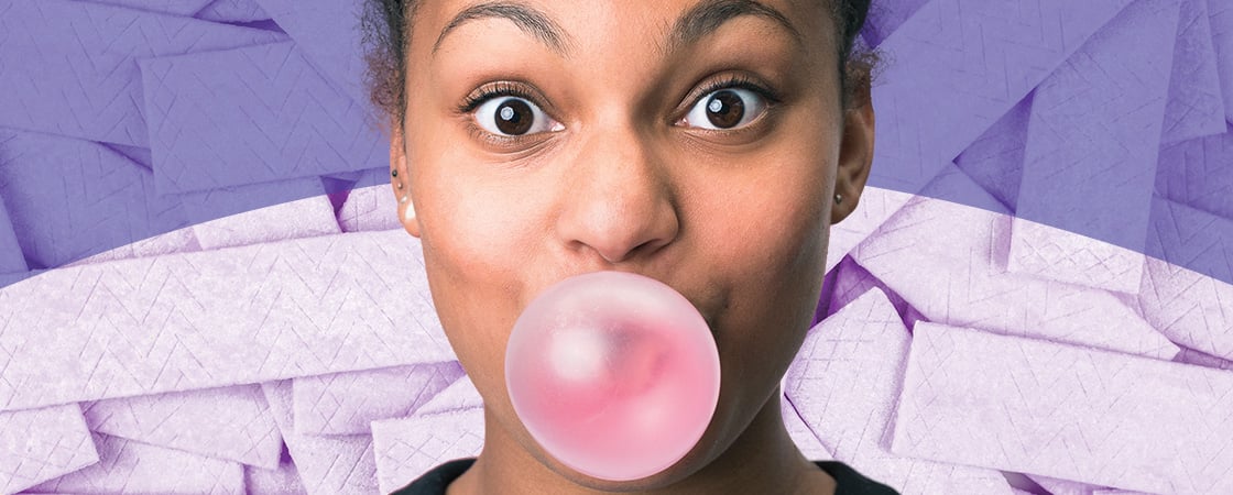 Image of a person blowing a bubble from pink gum and background is layers of purple sticks of gum