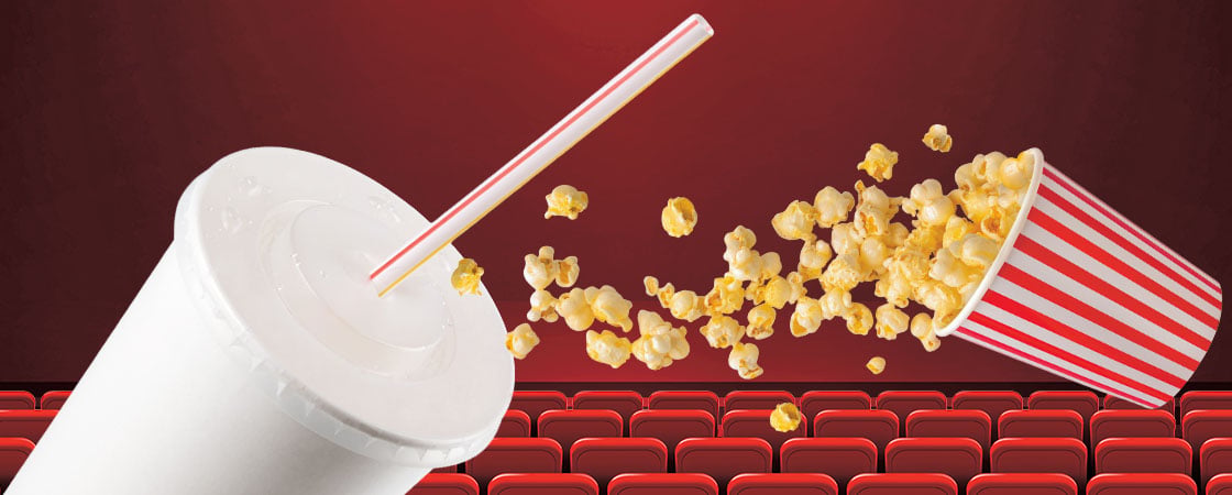 A fountain drink and a tipped bucket spill popcorn in front of movie theater seats