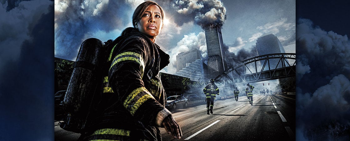 A firefighter stands on a city street as smoke rises from tall buildings in the background