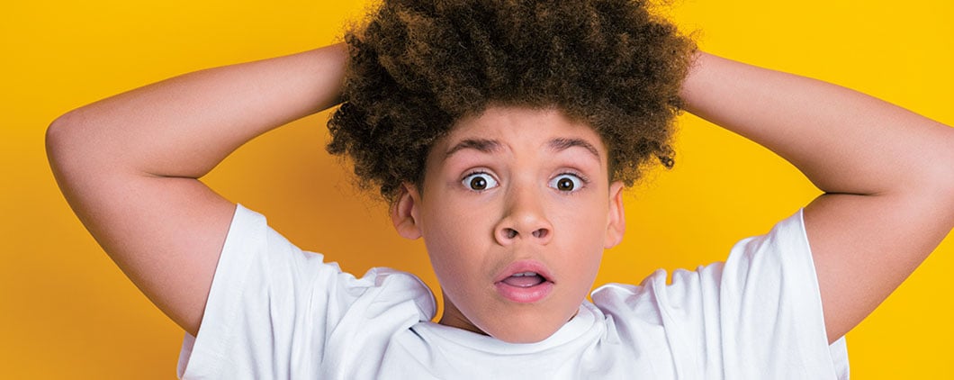 A boy holds both hands on top of his head with wide eyes and an open mouth