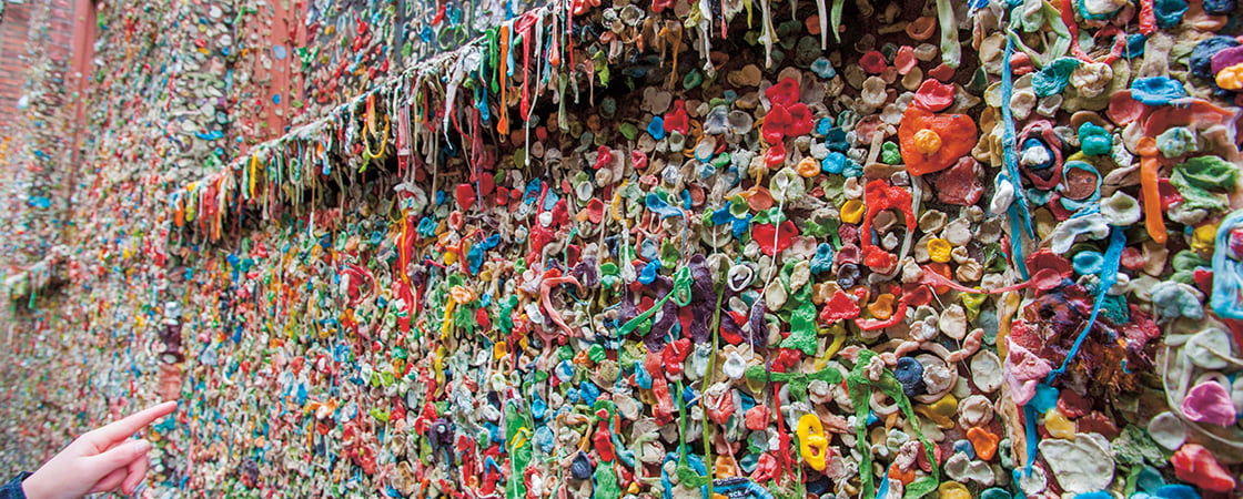 A brick wall covered in thousands of pieces of colorful chewing gum, with a hand visible at the left edge
