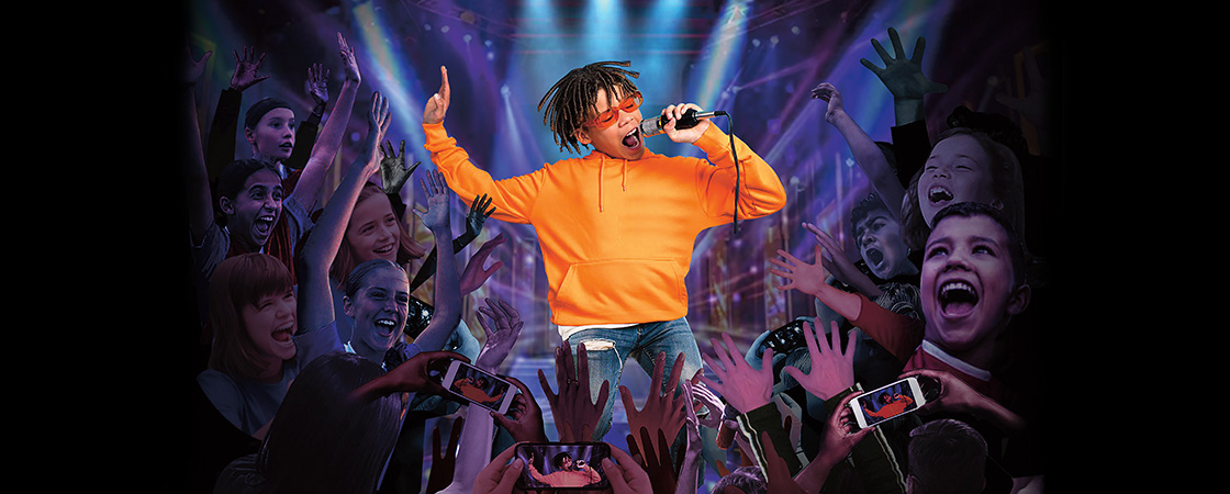 A boy performs on a lit stage with a microphone as a crowd of cheering children surrounds him