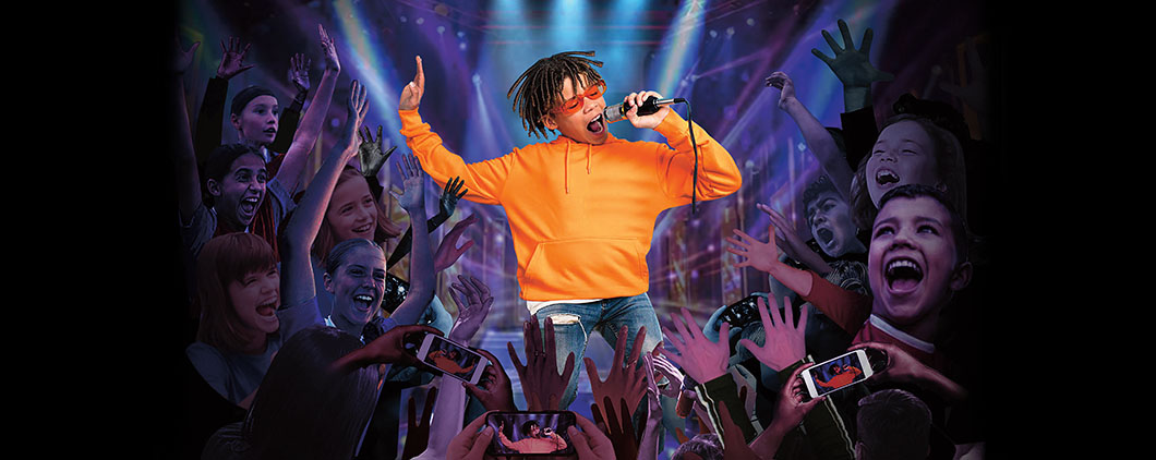 A boy sings into a microphone on a lit stage as a crowd of cheering children surrounds him
