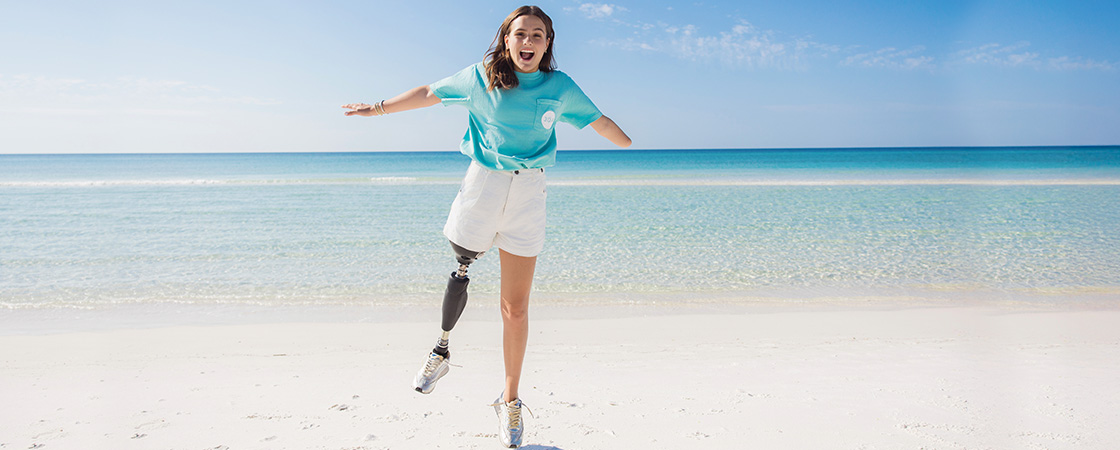 A young woman with a prosthetic leg jumps on a sandy beach with arms outstretched
